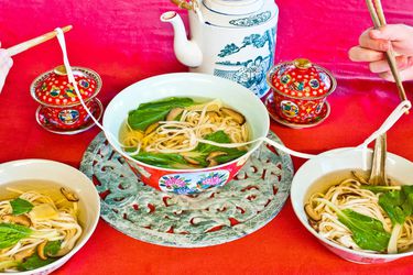 红桌子上放着一大碗长寿面，旁边放着一壶茶。食客们从碗里舀出面条，用筷子夹住。