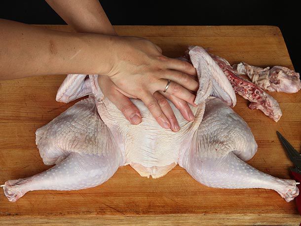 Two hands pressing firmly on the breast of a spatchcocked turkey in order to crack the breastbone and make the bird lie flat