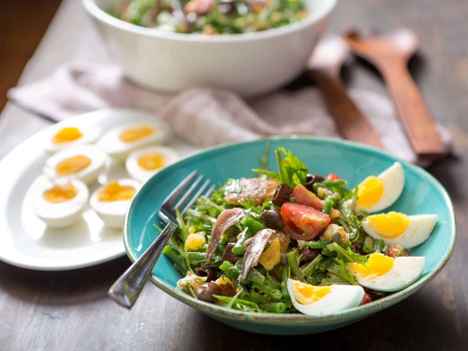 Nicoise salad on serving plate with eggs and salad in the background.