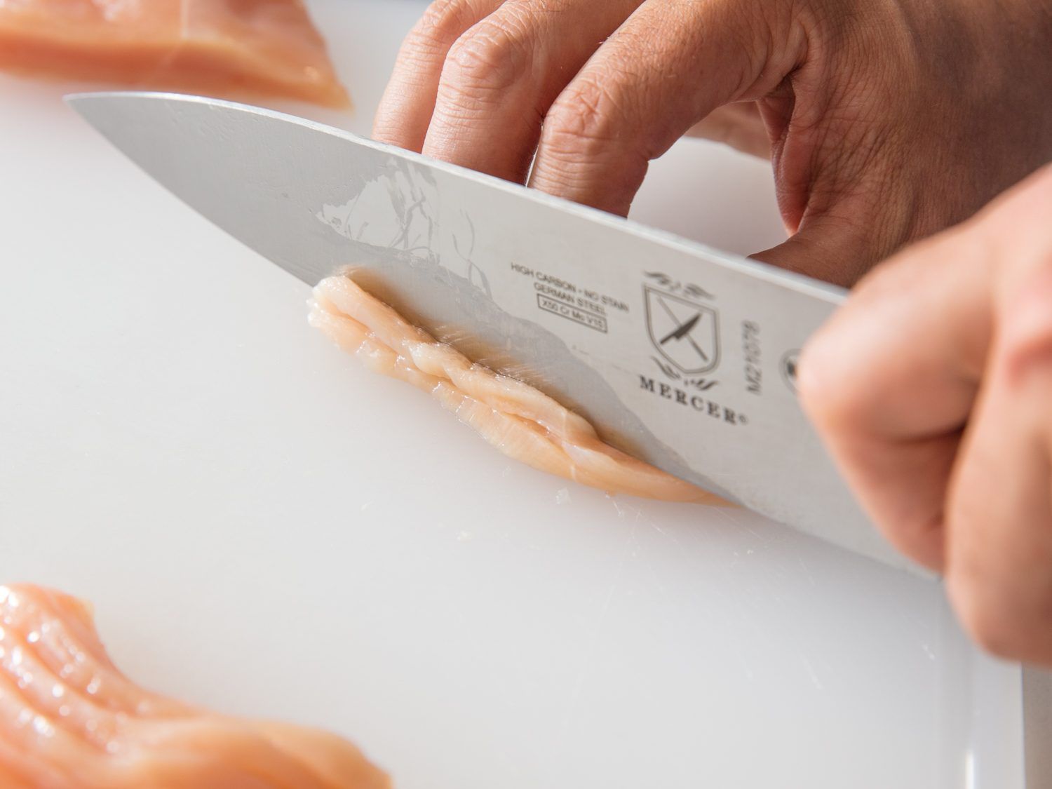 A knife slicing through two slices of chicken breast stacked on top of one another to create julienned strips for stir-frying.