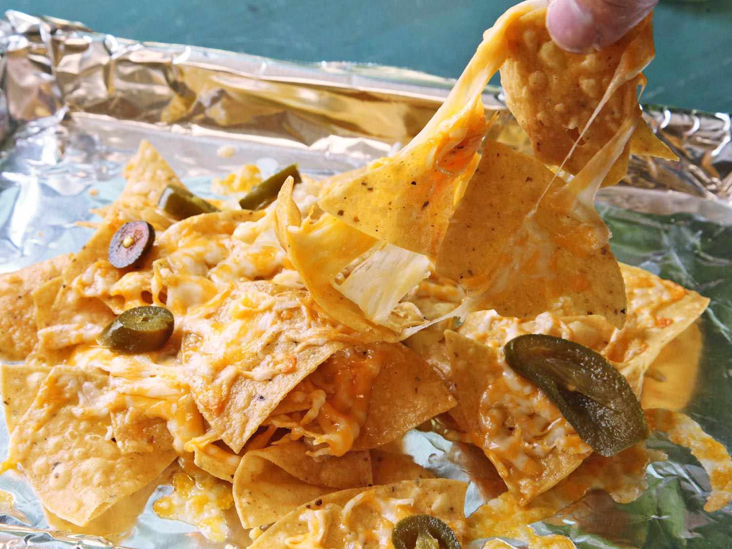 A cheese-encrusted chip is removed from a sheet pan of nachos, pulling several other chips with it.