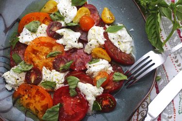 A caprese salad with heirloom tomatoes, mozzarella, and fresh basil on a plate with a fork and knife.
