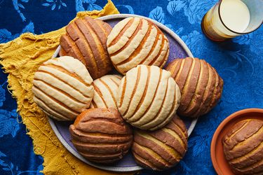 Overhead view of conchas on a plate with a glass of milk. Tablecloth is a velvety blue with a floral pattern and plate is resting on a bright yellow napking