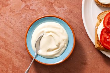 Two-minute mayonnaise in a serving bowl with a spoon, with an open face tomato sandwich on the right edge of the frame