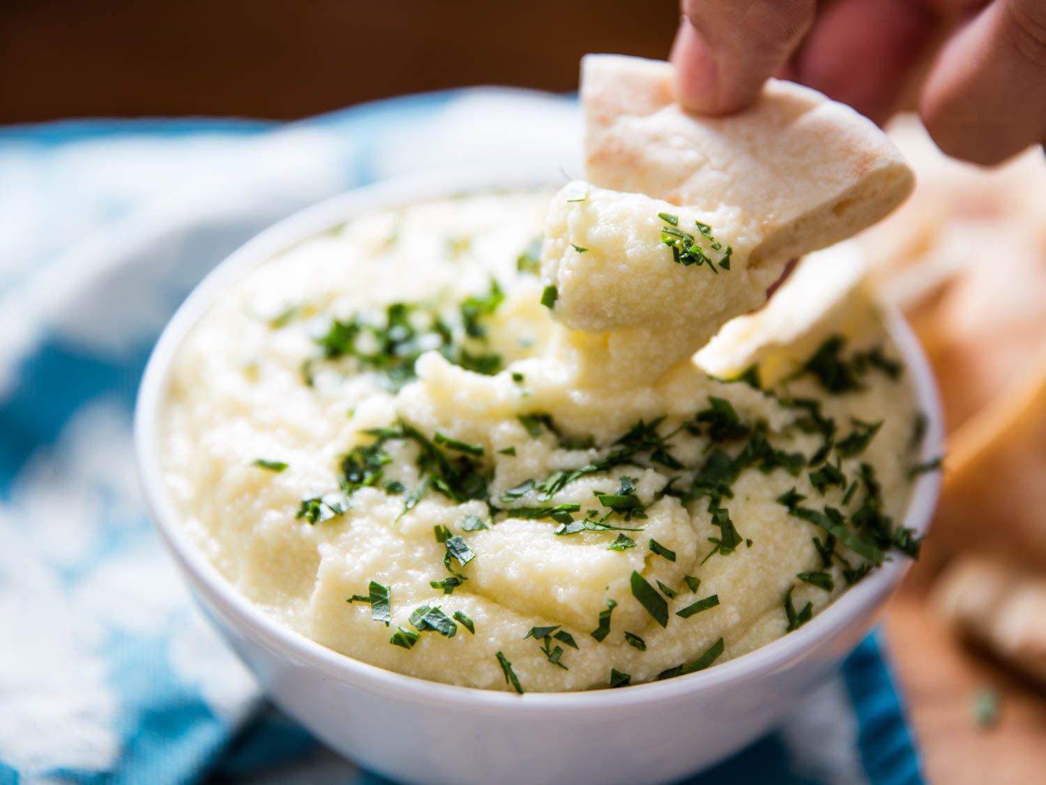 Hand holding pita slice that's being dipped into small bowl of potato puree topped with herbs.