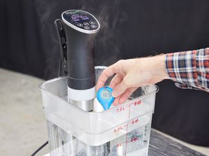 A sous vide machine heating up a cambro container filled with water and a hand with a thermometer taking the temperature of the water
