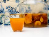 Side view of a glass of tepache next to a large batch in a gallon containter