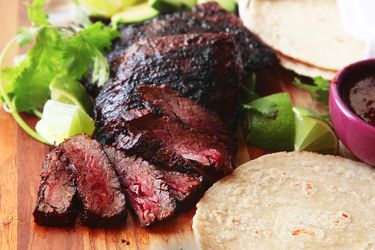 Sliced, grilled medium-rare carne asada fanned out on a wooden cutting board with corn tortillas, limes, and cilantro in the background.