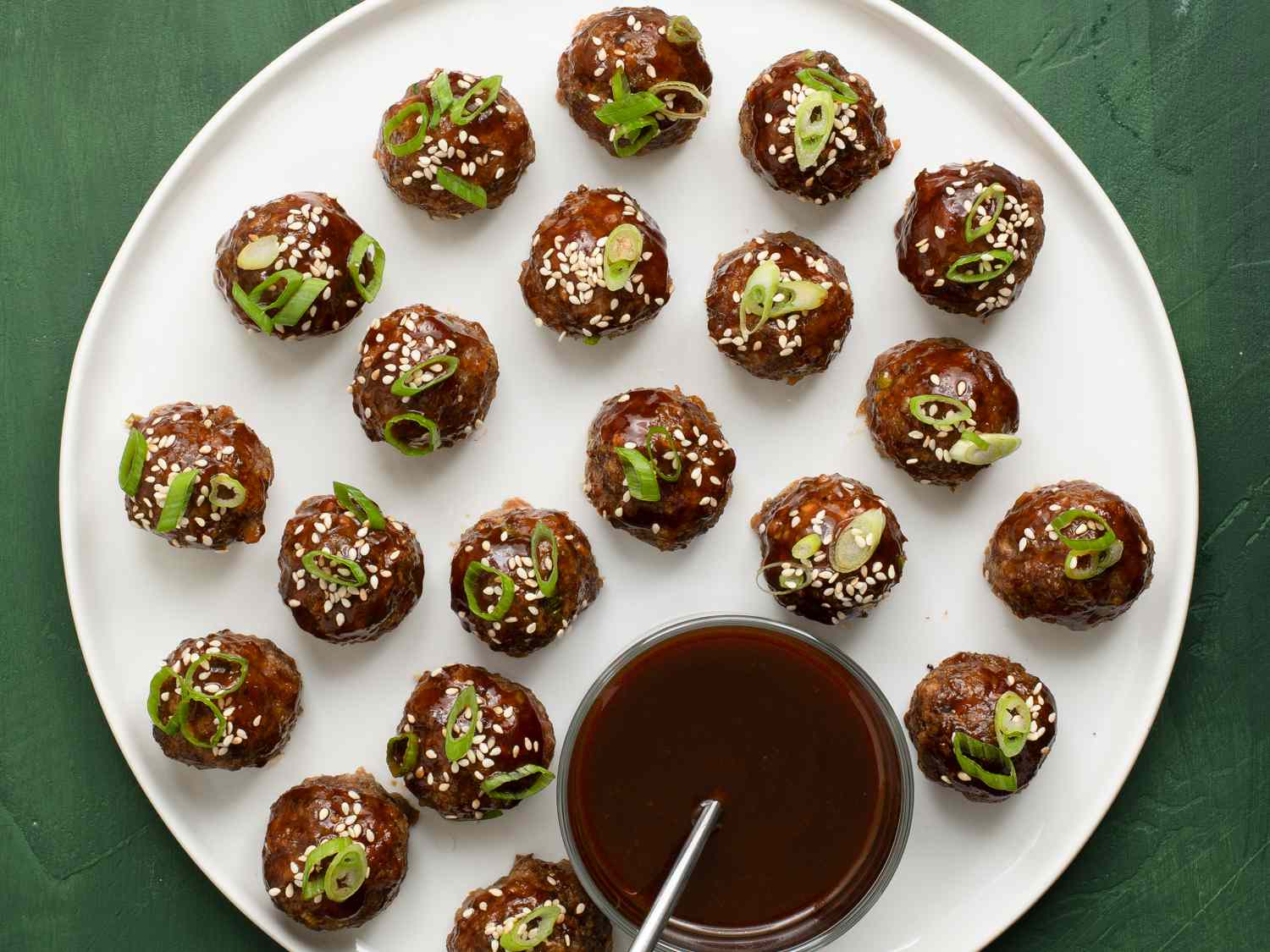 Hoisin-glazed cocktail meatballs on a white porcelain plate. There is a glass bowl of dipping sauce with a metal spoon on the plate.