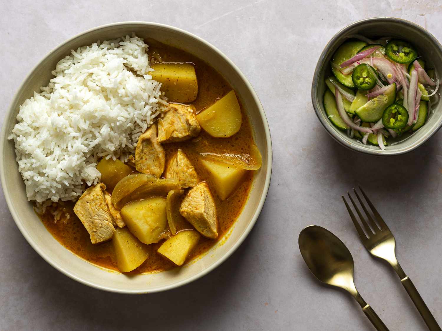A bowl of Thai chicken yellow curry in a cream colored ceramic bowl. On the right side of the image is a smaller bowl holding the accompanying cucumber salad. There are a fork and spoon in the bottom right hand corner of the image.