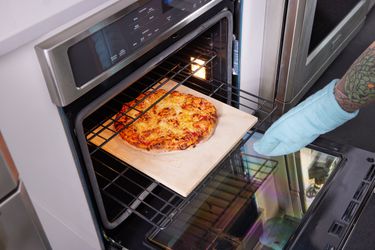 一只戴着烤箱手套的手把一块烤石从烤箱里拉出来，上面有一块披萨