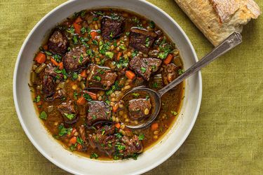 A white porcelain bowl holding beef barley soup. There's a metal spoon in the bowl and the bowl is palced on a green cloth. There is also part of a baguette in the top right corner of the image.