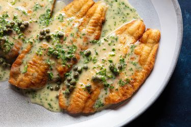 Deep golden brown fish fillets on a large serving platter are covered in a creamy, glossy, perfectly emulsified butter sauce, studded with capers and flecked with minced parsley.