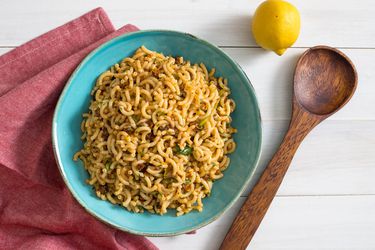 A bowl of pasta salad with Spanish chorizo and piquillo peppers. A lemon, wooden spoon, and red kitchen towel are next to the bowl.