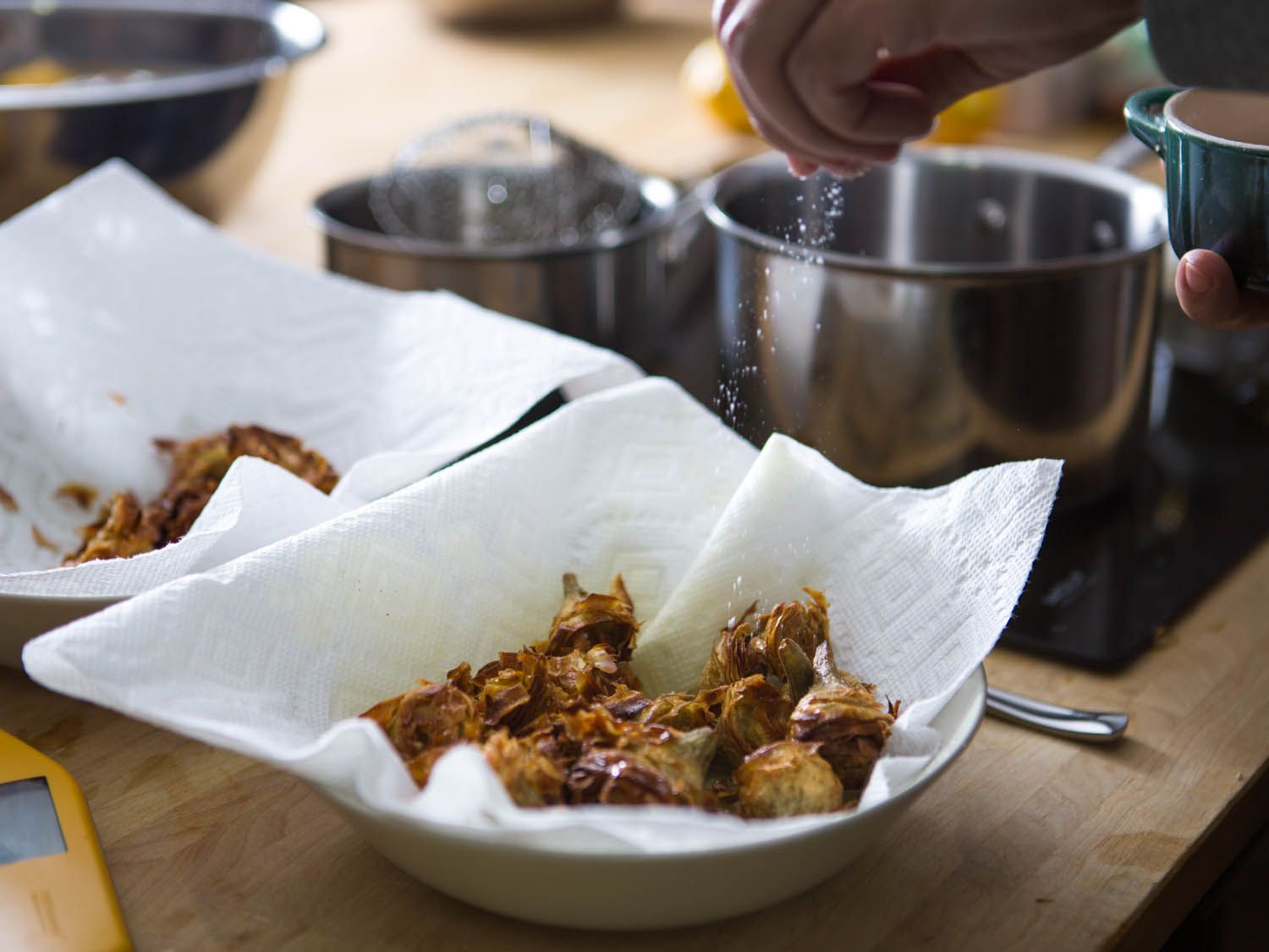 Sprinkling salt on fried artichoke hearts drained on paper towel-lined bowls.