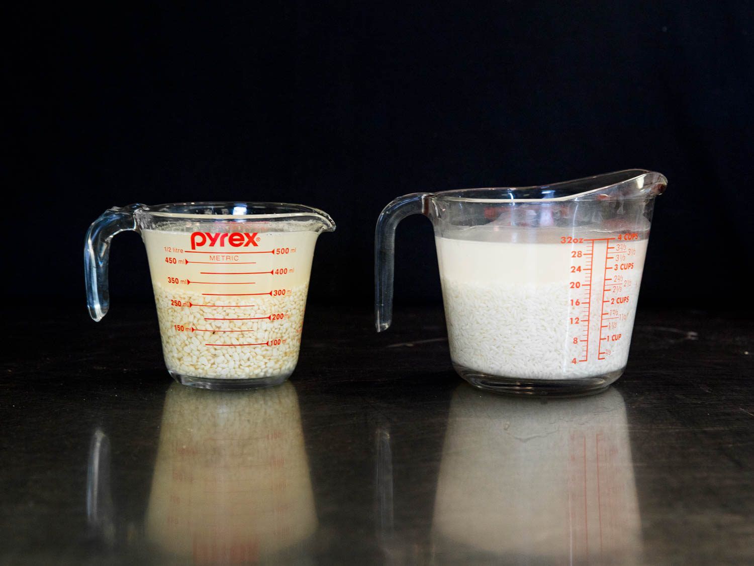 Two glass measuring cups holding grains of rice and urad dal soaking in liquid.