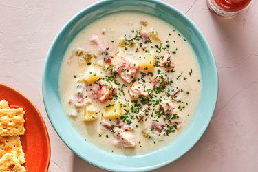 A baby blue bowl holding a serving of creamy salmon chowder. The chowder is topped with minced herbs and there is a red plate of saltine crackers in the bottom left corner of the image, and a bottle of Tabasco hot sauce in the top right corner of the image.