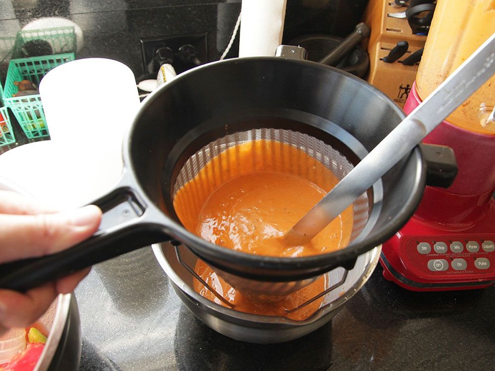 Pressing blended gazpacho through a fine-mesh strainer with a ladle.