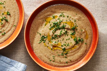 An orange-brown ceramic bowl holding creamy mushroom soup topped with chopped herbs and drizzled olive oil.