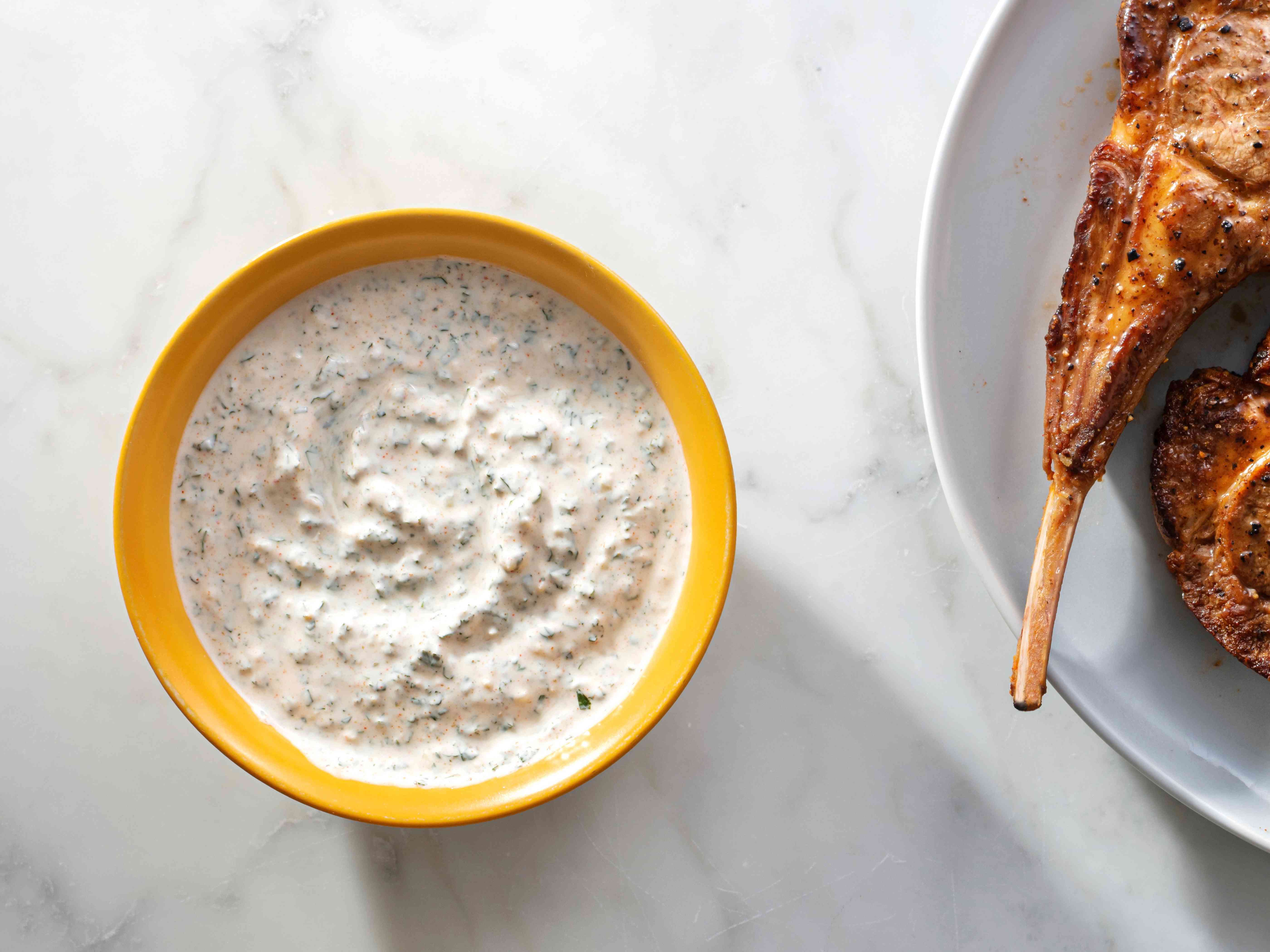 Yogurt mint sauce in a yellow bowl on a marble background, with lamb chops on a plate on the right edge of the photo.