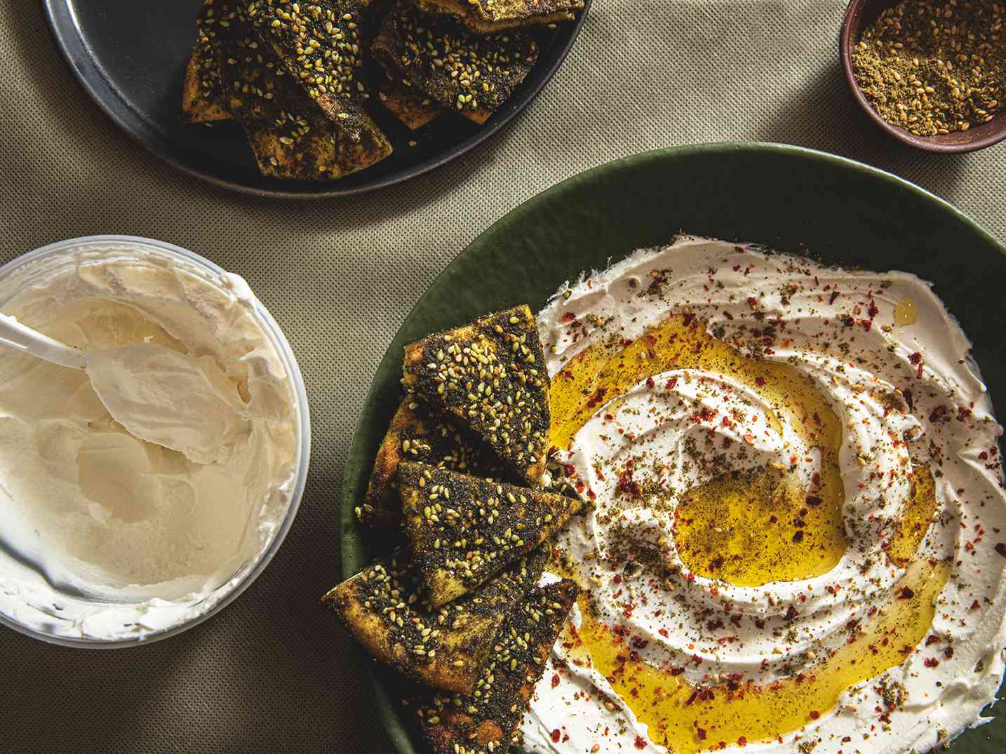 Overhead view of a plate of labneh with zatar pita chips surrounded by a grocery store container of labneh, a bowl of zatar, and a plate of pita chips