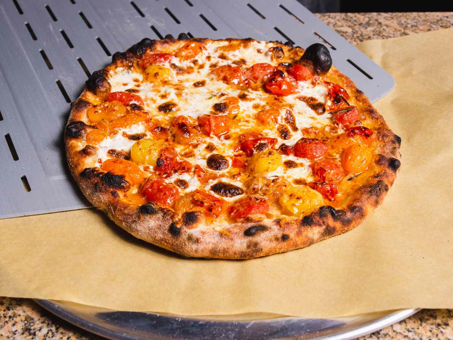 Burst cherry tomato outdoor oven pizza being slid off a peel onto parchment on a pizza pan