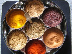 A collection of seven dry spices in a masala dabba, a large tin with smaller tins inside.