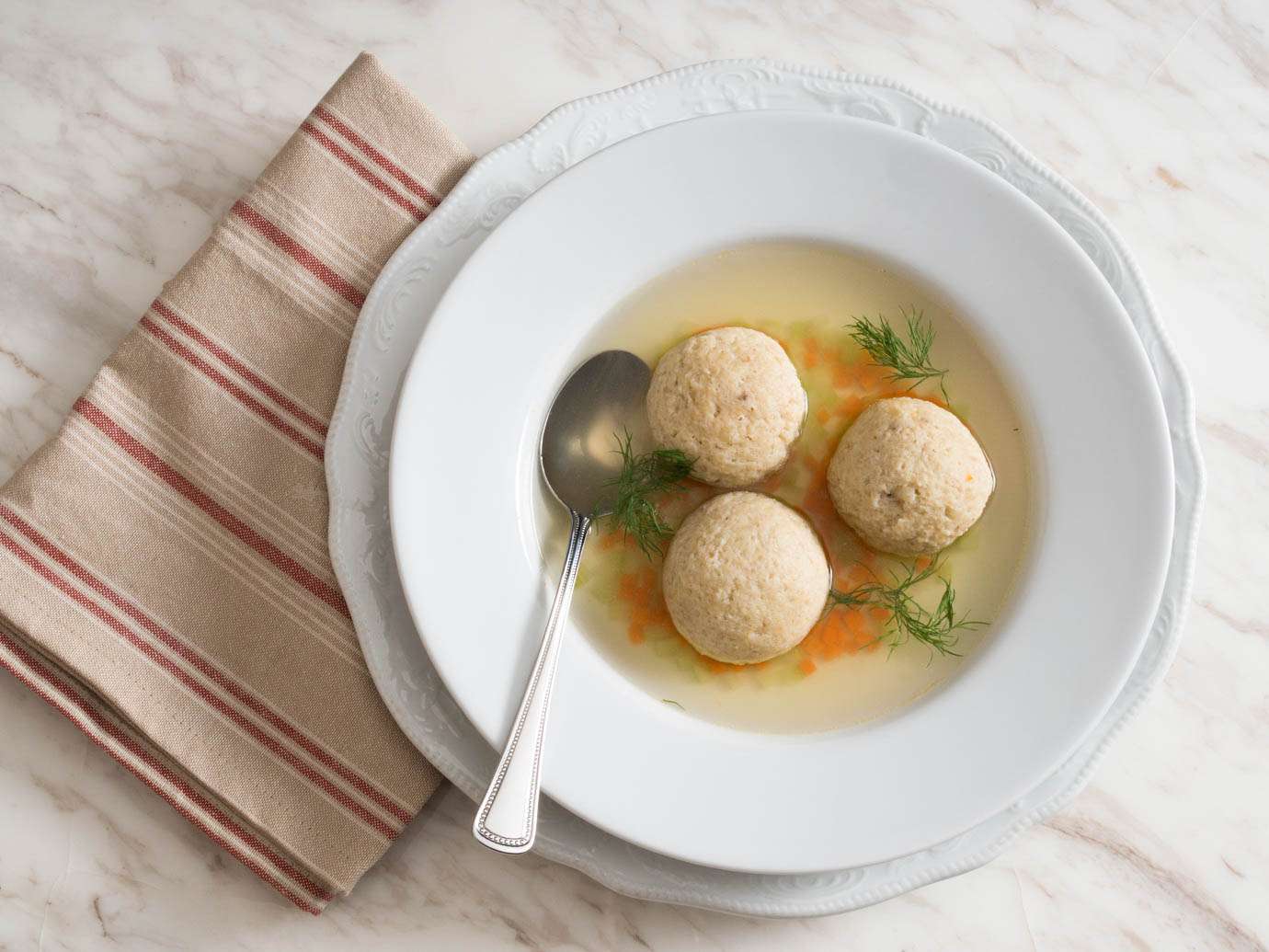 An overhead shot of a broad-rimmed white soup bowl containing three matzo balls in a light broth. Finely diced carrot and celery have sunk between the balls, and several dill fronds are floating on the surface.