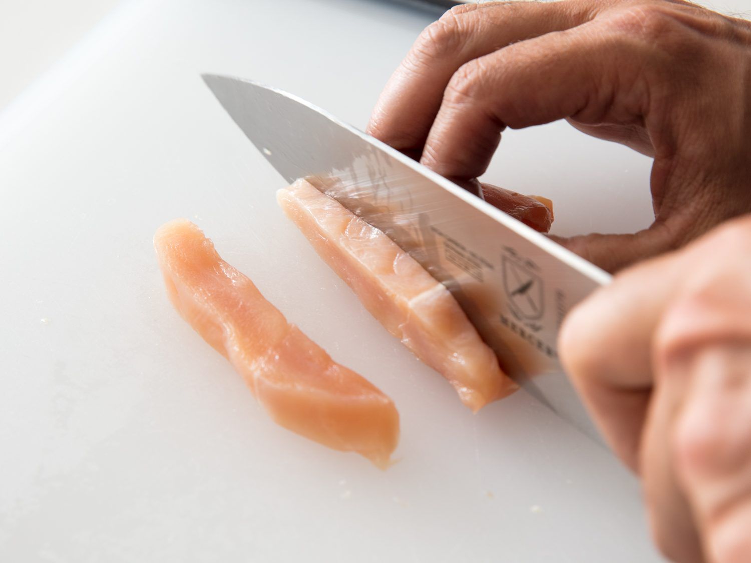 A knife slicing against the muscle grain of a chicken breast to create larger slices, which in turn will be diced into cubes, for stir-frying.
