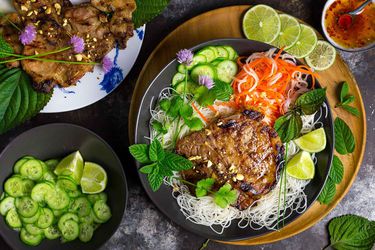 Overhead shot of a bowl of vermicelli noodles topped with grilled pork chops, carrot-daikon pickles, cucumbers, perilla/shiso, mint, cilantro, crushed peanuts, and a few stray flowering chives. A small bowl of nuoc cham is close at hand, as well as a perilla-leaf-lined plate piled with more of the grilled pork chops and a bowl of sliced cucumbers and lime wedges.