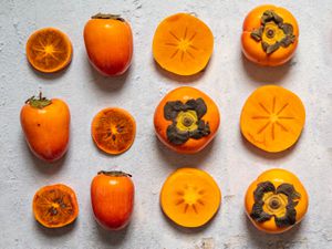 A variety of persimmons, some cut and some whole, arranged in a grid on a stone surface