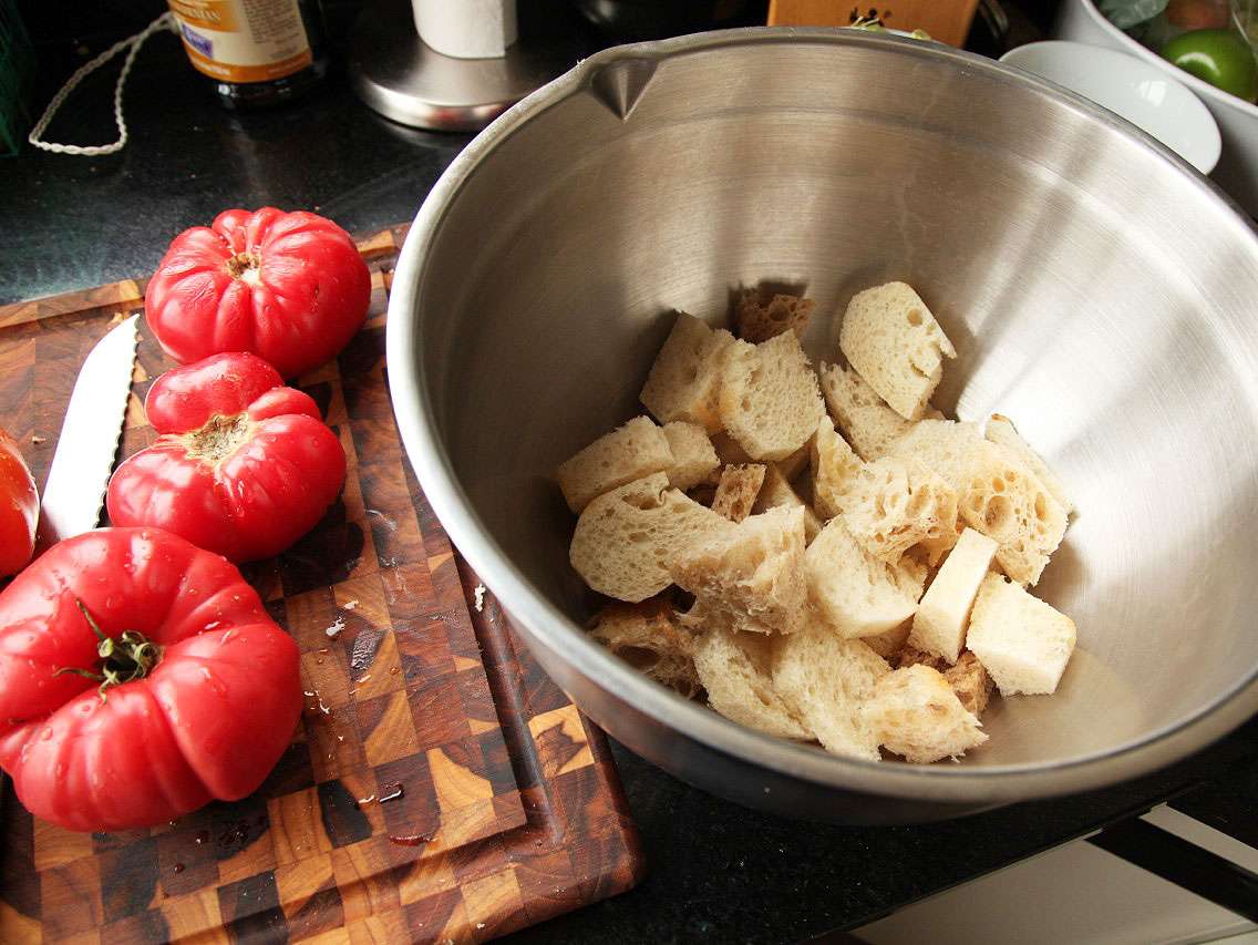 Pieces of torn bread with crusts removed, in a large mixing bowl next to several tomatoes lying on a cutting board.