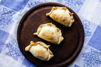 3 empanadas on a wooden plate on top of a traditional Argentinian fabric with a blue and white checkered pattern