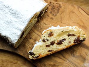 Buttery marzipan stollen dusted with powdered sugar on a wood cutting board. A slice of stollen is laying on the cutting board, exposing the fruit and marzipan inside.