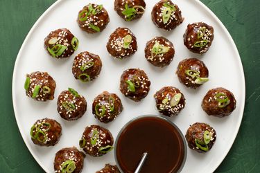 Hoisin-glazed cocktail meatballs on a white porcelain plate. There is a glass bowl of dipping sauce with a metal spoon on the plate.