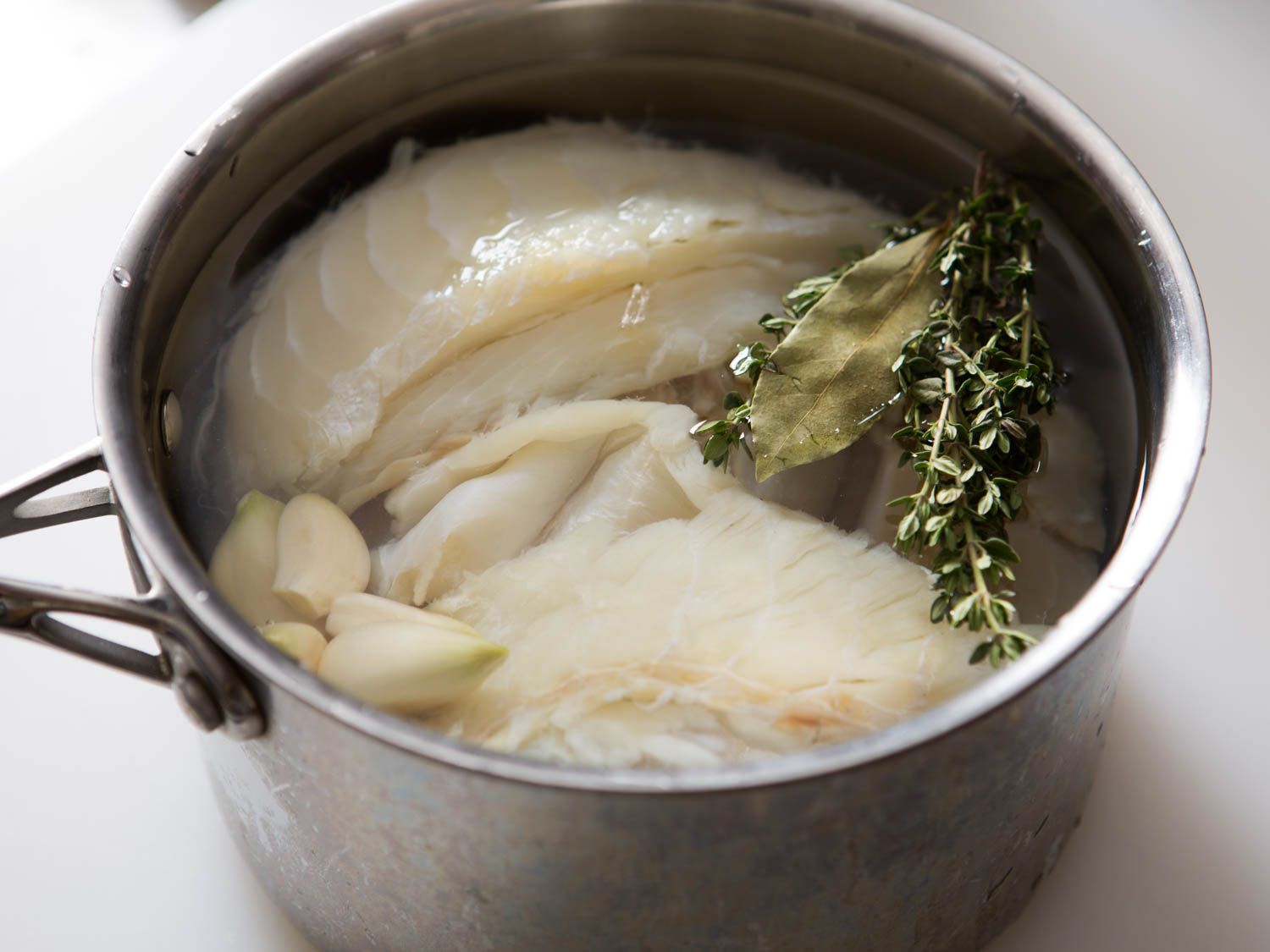 一个不锈钢锅，里面有盐鳕鱼，水，大蒜，新鲜的百里香和一片干月桂叶。
