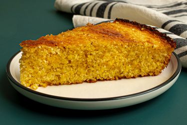 A slice of Southern-style unsweetened cornbread on a round white ceramic plate with a dark green rim. The cornbread is resting on a dark teal surface and there's a dishcloth in the background.