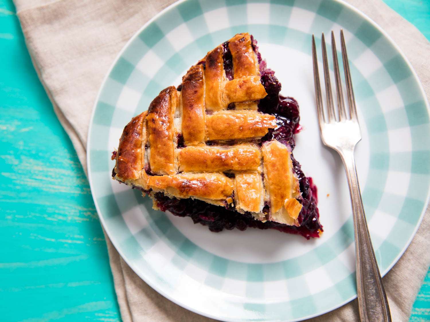 一个楔形人字形的蓝莓派，上面有格子，放在格子边的盘子上，用叉子叉着