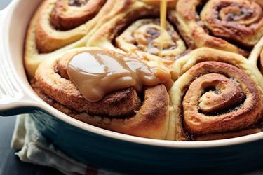 A ceramic baking dish with cinnamon sticky buns. Glaze is being drizzled over them.