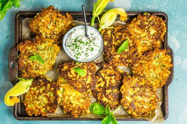 Overhead view of a sheetpan piled high with zucchini-and-corn fritters and a ramekin of herb sour cream. The fritters are garnished with squash blossoms and basil sprigs.