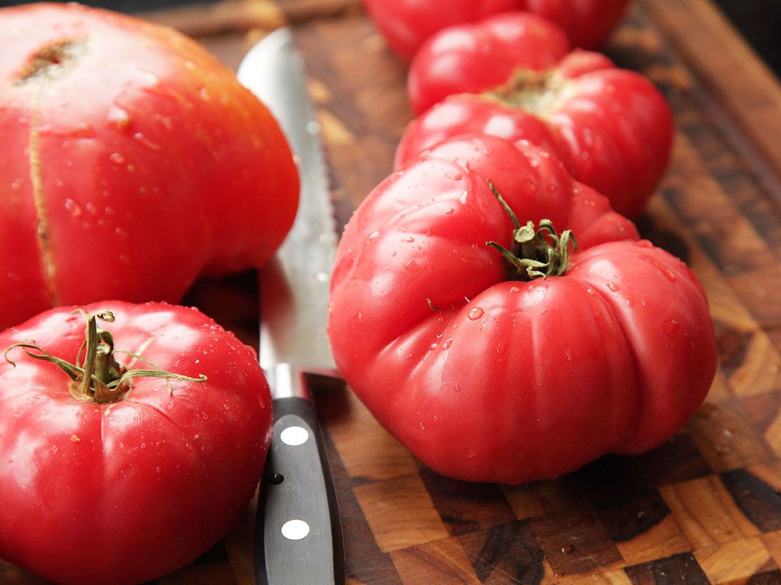 A close up of several tomatoes for gazpacho and a knife on a wooden cutting board.