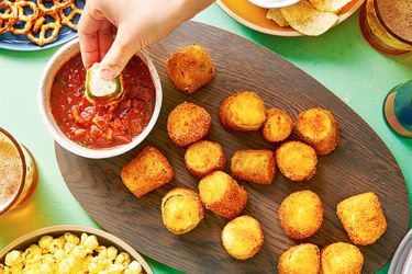 An oval wooden platter holding crispy deep-fried jalapeÃ±o poppers and a small bowl of tomato sauce. There is a hand dipping a popper into the tomato sauce, and the periphery of the image contains other snacks, such as pretzels, popcorn, and chips.