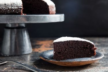 A slice of dairy-free chocolate cake served on a slate-colored plate. The remaining cake is nearby, perched on a stainless steel cake stand.