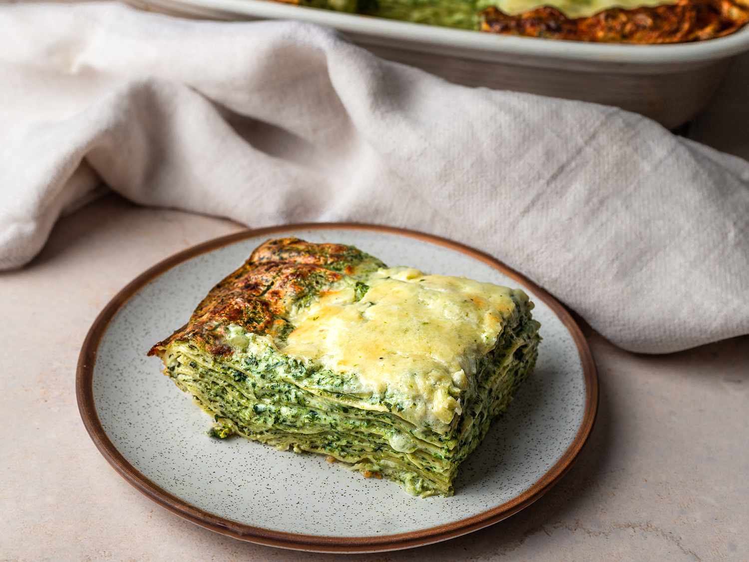 A slice of spinach lasagna on a ceramic plate, with a cloth and the remaining lasagna behind it.