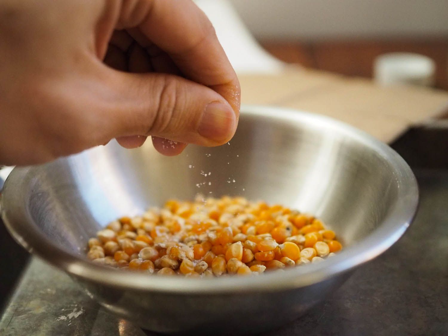 一个小的金属搅拌碗，里面装着黄色的爆米花仁和油，一只手在碗上撒上粗盐。