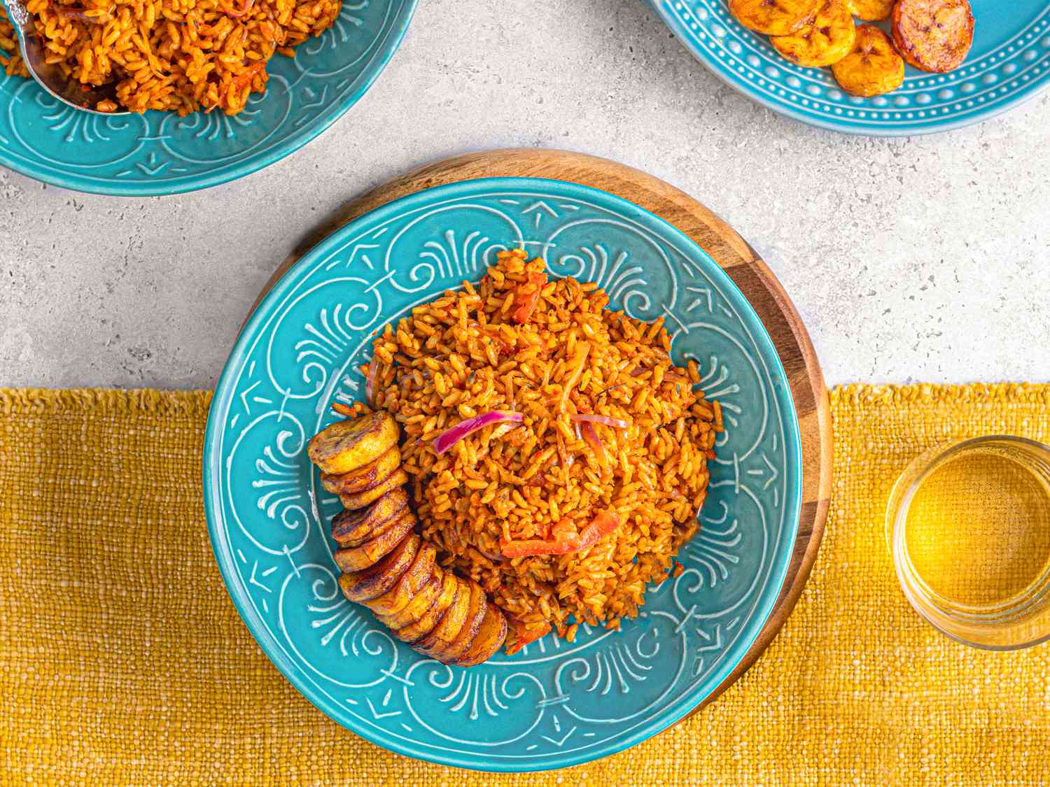 Overhead view of Jollof rice on a bright blue plate on top of a yellow table runner next to a glass of water.