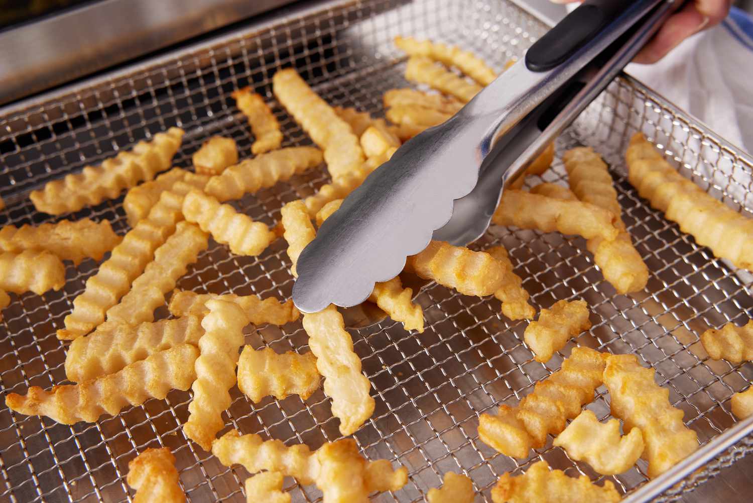 A person using tongs to remove cooked fries from an air fryer toaster oven's basket