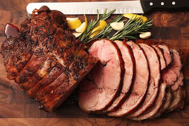 Slow-roasted boneless lamb leg on a cutting board. Some of it is sliced. There are fresh rosemary sprigs, whole garlic cloves, and lemon wedges next to it.