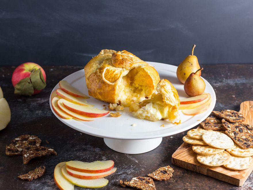 Baked brie in a pastry shell with apple and pear compote, surrounded by thinly sliced apples, crackers, and whole fruit.
