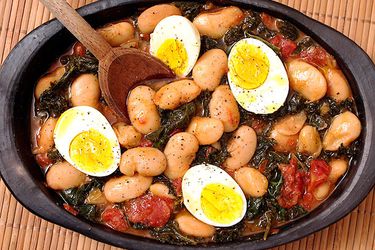 Overhead view of butter beans with kale and eggs, served in a black clay platter.